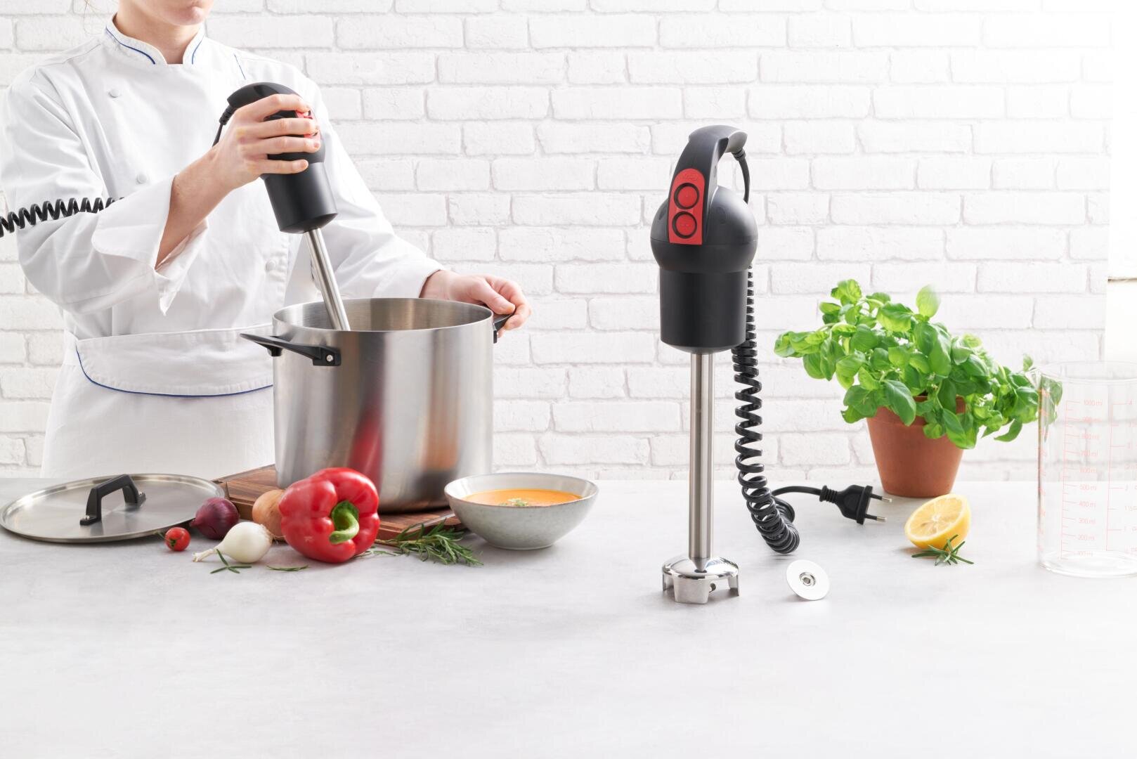 Ein Koch in weißer Uniform verwendet einen Stabmixer in einem großen Topf auf einer Küchentheke. Daneben stehen Kräuter, Gemüse, eine Schüssel Suppe, ein zweiter Mixer und eine Basilikumpflanze im Topf.