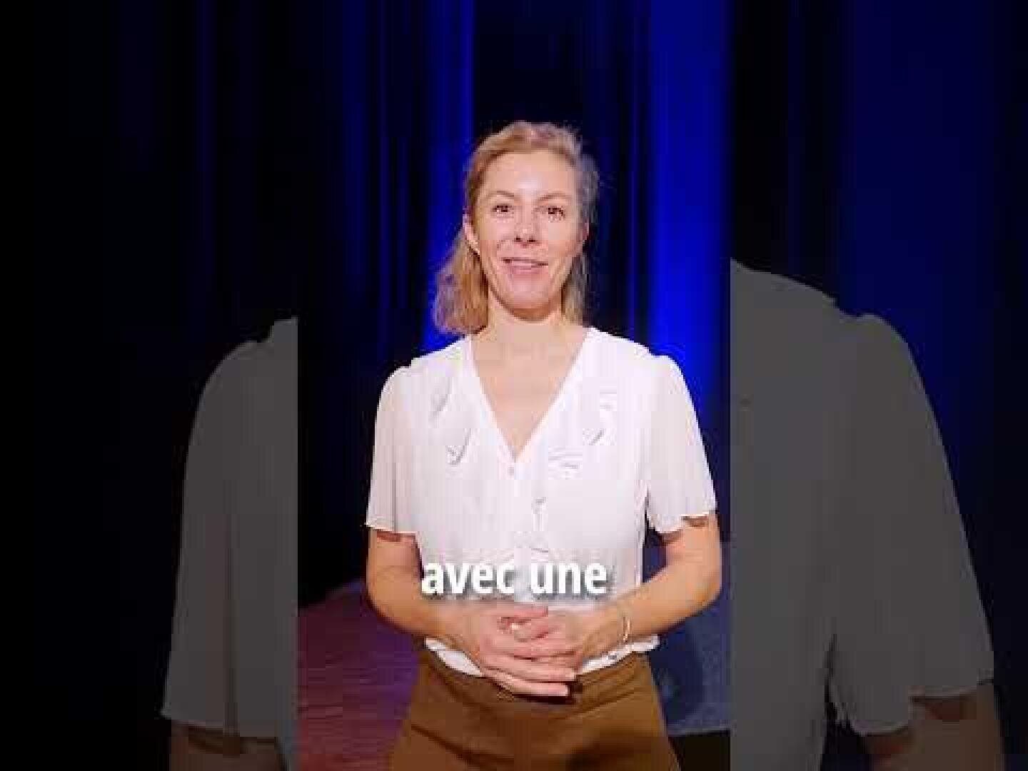 Une femme aux cheveux blonds, vêtue d'un chemisier blanc et d'un pantalon marron, se tient sur une scène à l'arrière-plan bleu foncé. Le texte français avec une apparaît dans la partie inférieure de l'image.