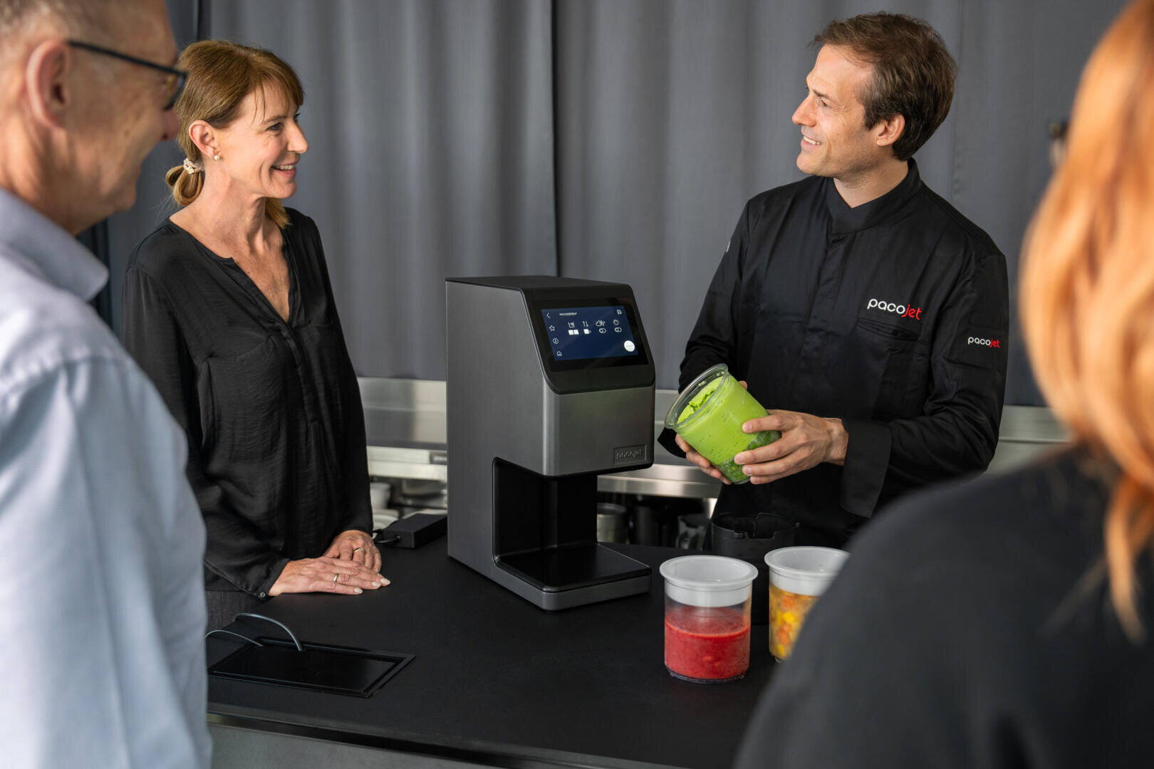 Un homme vêtu d'une blouse noire de chef cuisinier fait la démonstration d'un appareil de cuisine moderne à trois personnes. Il tient un récipient contenant des aliments verts, tandis que des bacs contenant des ingrédients rouges, jaunes et orange sont posés sur le comptoir.