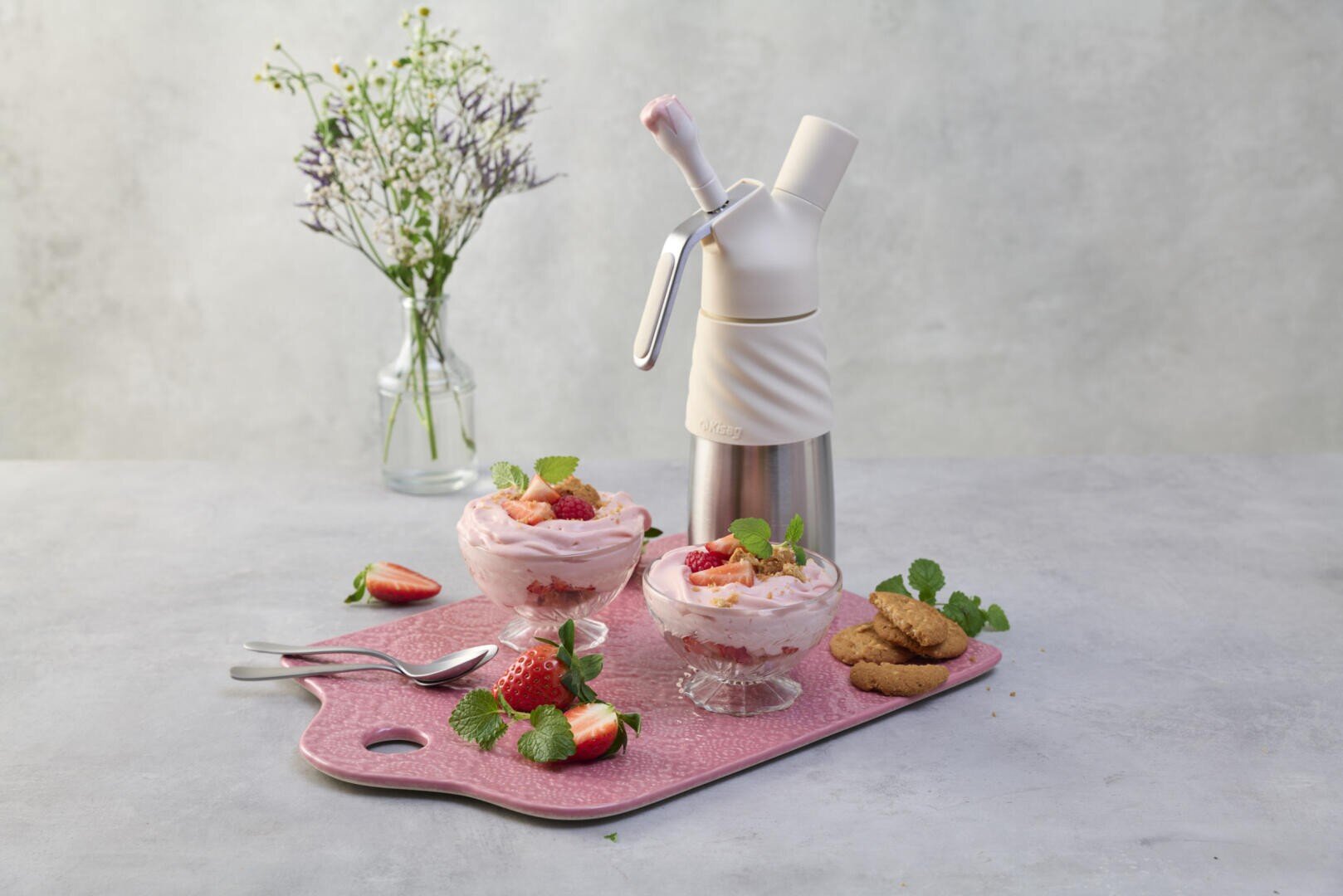 Deux coupes en verre contenant de la mousse à la fraise, garnie de menthe et de fraises, sont posées sur un plateau rose orné de biscuits et de fraises. À l'arrière-plan, on aperçoit un siphon à chantilly, des cuillères et un vase fleuri.