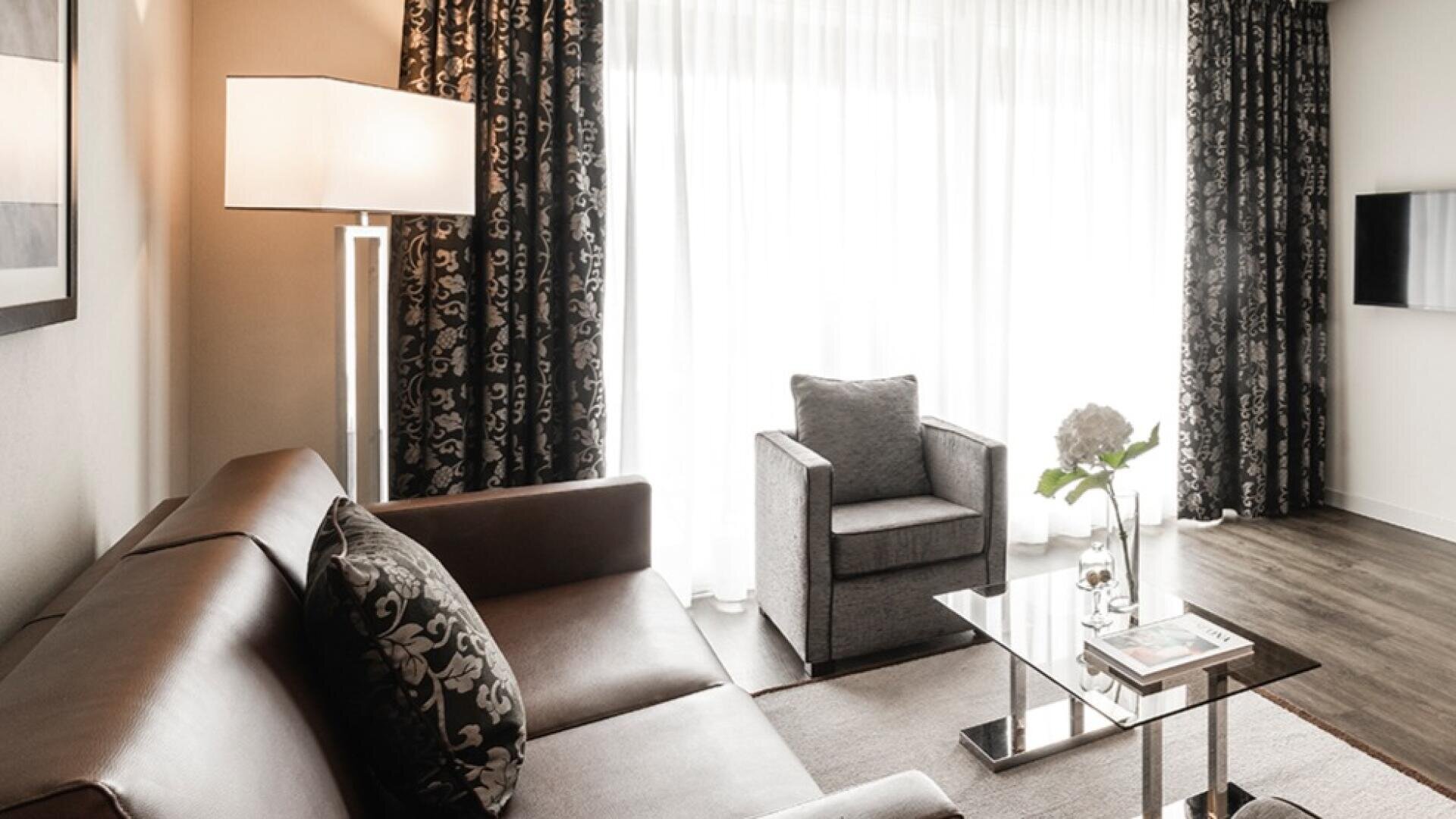 Modernes Wohnzimmer mit einem braunen Sofa, gemusterten Kissen, einem grauen Sessel, einer Stehlampe, einem Couchtisch aus Glas mit einer Blumenvase, einer Zeitschrift und einem großen Fenster mit durchsichtigen und dunkel gemusterten Vorhängen.