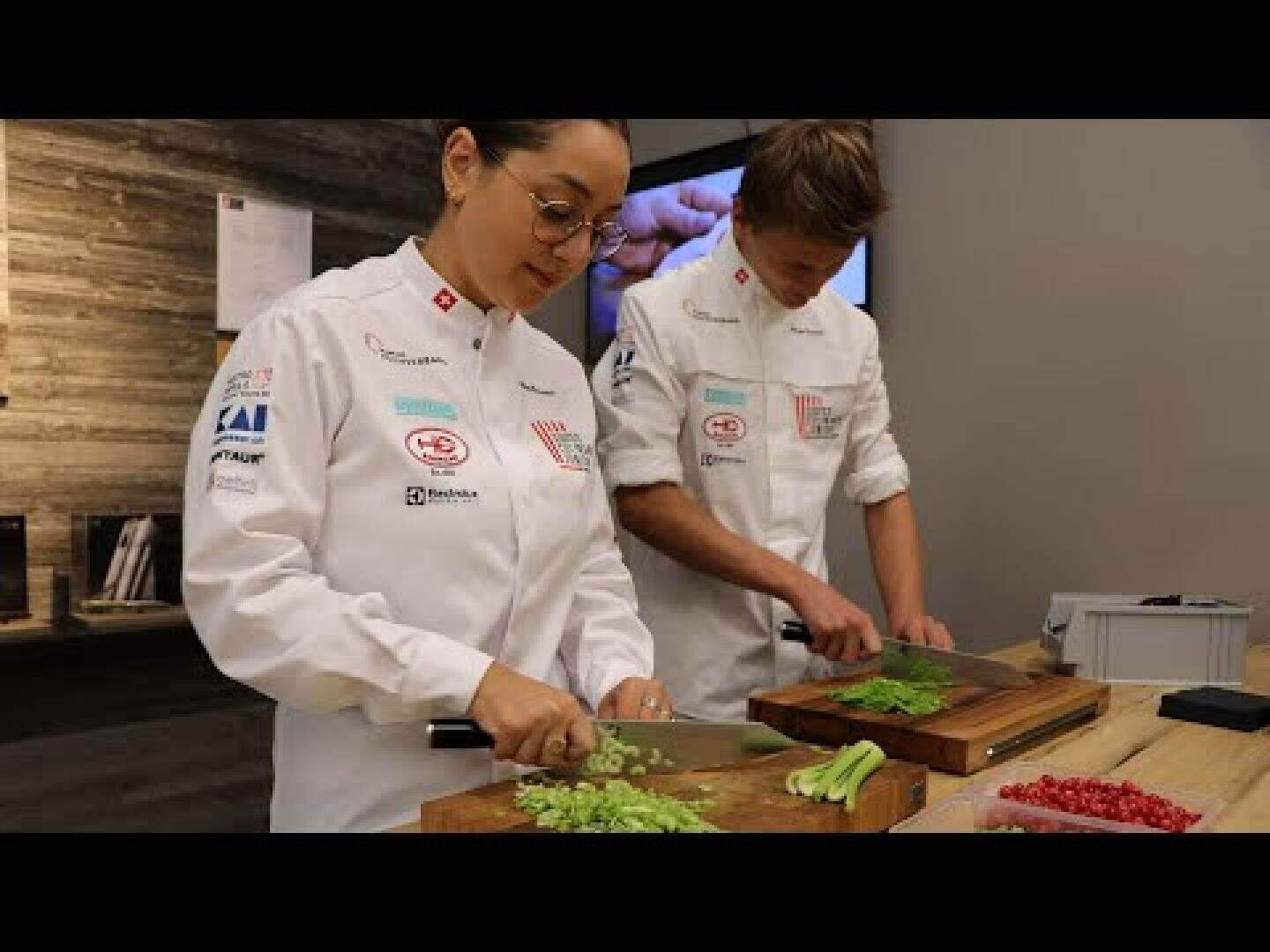 Deux cuisiniers en uniforme blanc hachent des légumes verts sur des planches à découper en bois dans une cuisine moderne. Sur le comptoir, on peut voir différents ustensiles de cuisine et des ingrédients.