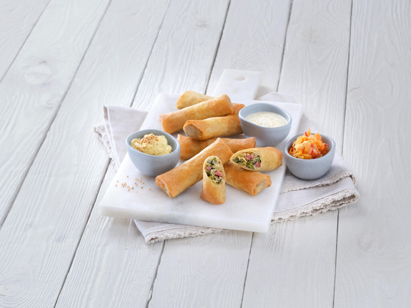Sur une table en bois blanc, une planche en marbre présente des rouleaux de printemps croustillants, dont certains sont ouverts pour laisser apparaître une garniture de légumes, accompagnés de deux sauces de trempage et d'un petit bol de légumes marinés.