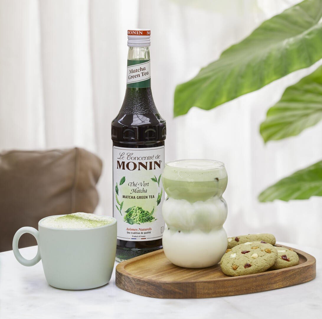 Eine Flasche Monin Matcha Green Tea steht hinter einem grünen Matcha-Latte in einem Becher, einem eisgekühlten Matcha-Latte in einem runden Glas und drei Keksen auf einem Holztablett, alles auf einem weißen Tisch mit grünen Blättern im Hintergrund.
