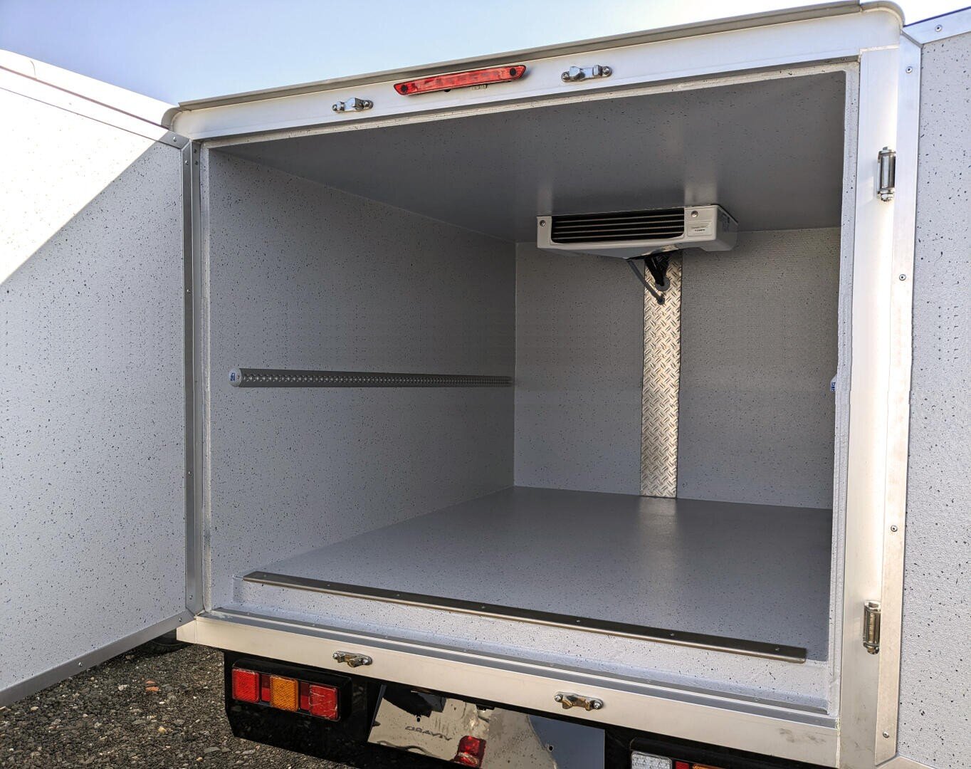 Vue arrière ouverte d'un compartiment de camion frigorifique vide, montrant des parois isolées propres et gris clair, une unité de refroidissement montée au plafond et une bande métallique sur la paroi gauche pour fixer la cargaison.