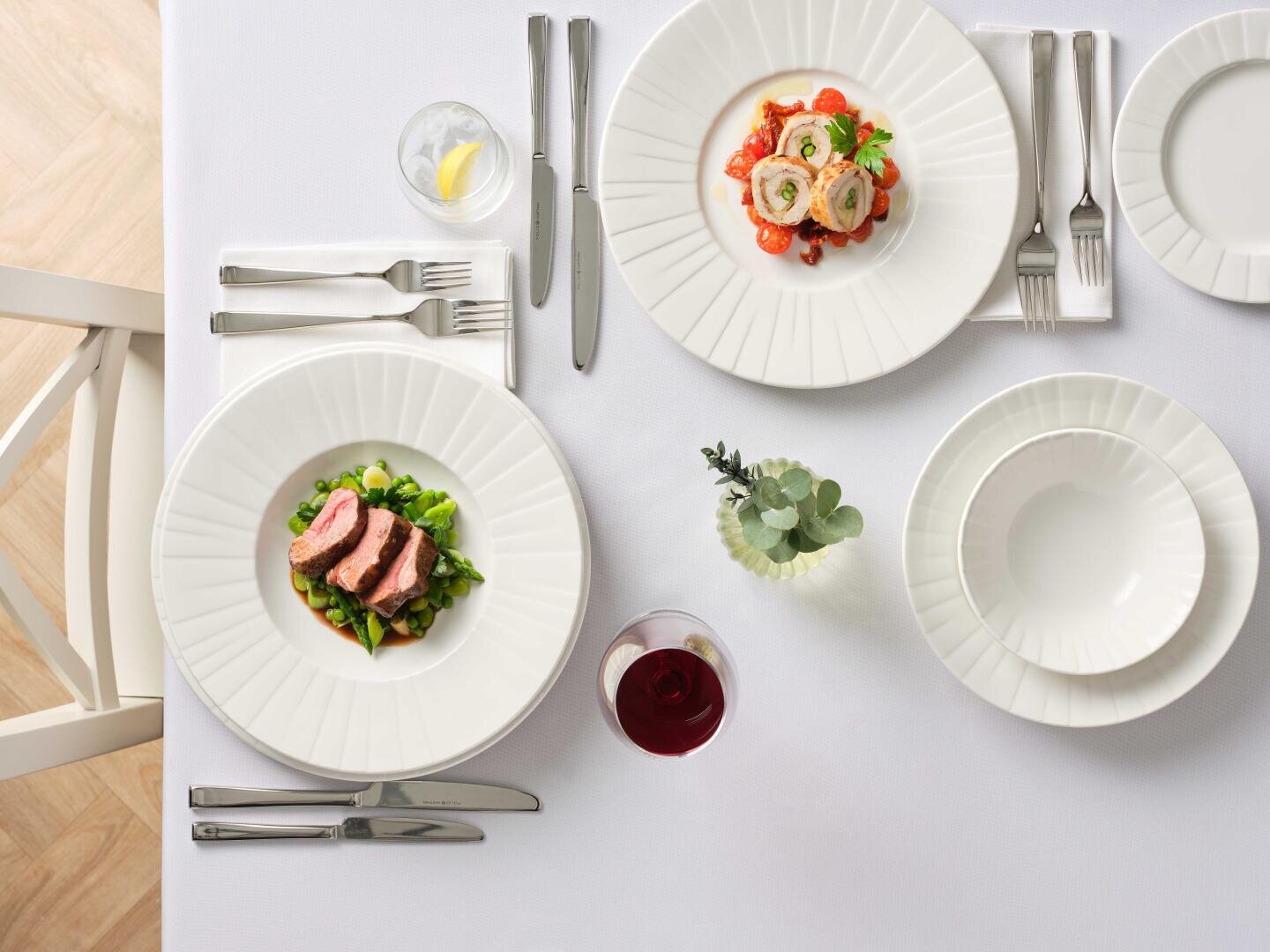 Une table blanche soigneusement dressée avec deux plats gastronomiques, un verre de vin rouge, un verre d'eau citronnée, des bols vides et des couverts en argent. Au milieu se trouve un petit vase avec de la verdure.