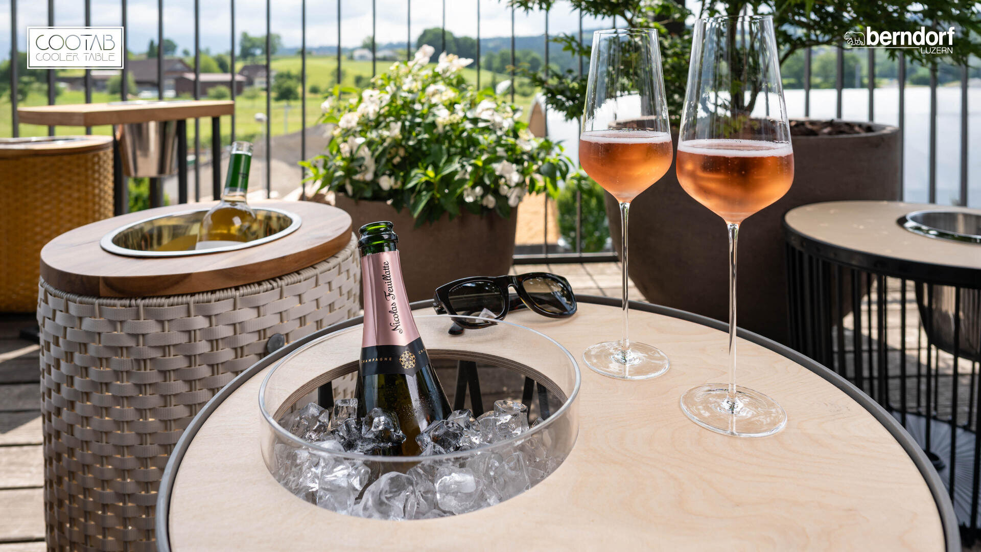 Une table ronde d'extérieur avec un seau à glace intégré contenant une bouteille de rosé pétillant, à côté de deux verres à vin remplis et de lunettes de soleil, installée sur un patio avec vue sur la verdure et un lac.