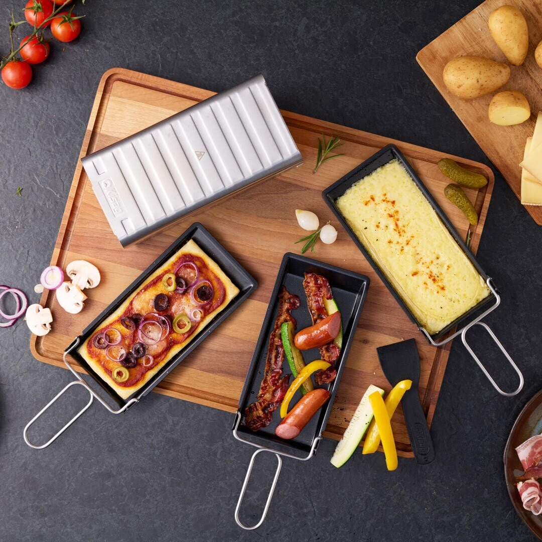 Ein Holzbrett mit einem Käseschmelzer, zwei Raclette-Pfännchen - eines mit geschmolzenem Käse, das andere mit Pizza und Zwiebeln - und ein weiteres Pfännchen mit Würstchen, Gemüse, Kartoffeln, Käsescheiben, Pilzen, Zwiebeln, Tomaten und Essiggurken in der Nähe.