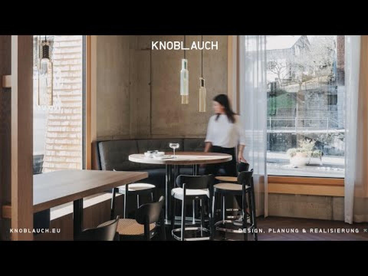 Eine Frau steht an einem runden Tisch mit Stühlen in einem modernen, minimalistischen Essbereich mit großen Fenstern, Betonwänden und hängenden Pendelleuchten. Die Worte KNOBLAUCH und DESIGN, PLANUNG & REALISIERUNG sind sichtbar.