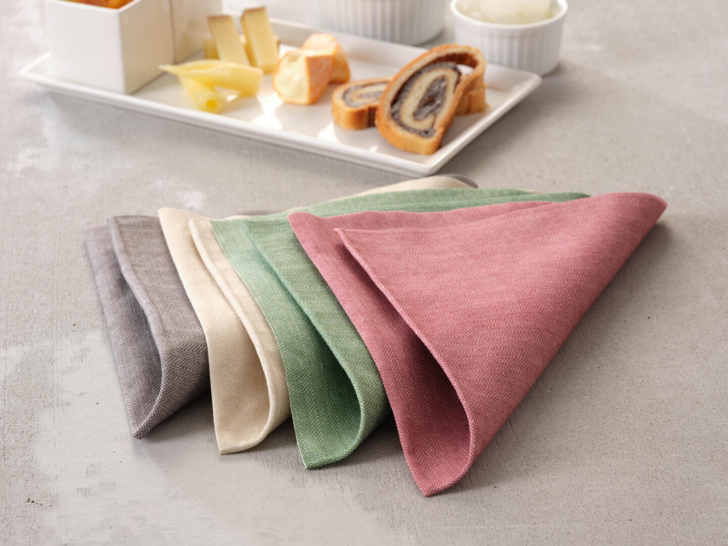 Vier fein säuberlich gefaltete Stoffservietten in Grau, Beige, Grün und Rosa sind in einer Reihe auf einer hellen Fläche angeordnet. Im Hintergrund steht ein weißes Tablett mit Käse, Gebäck und anderen Snacks.