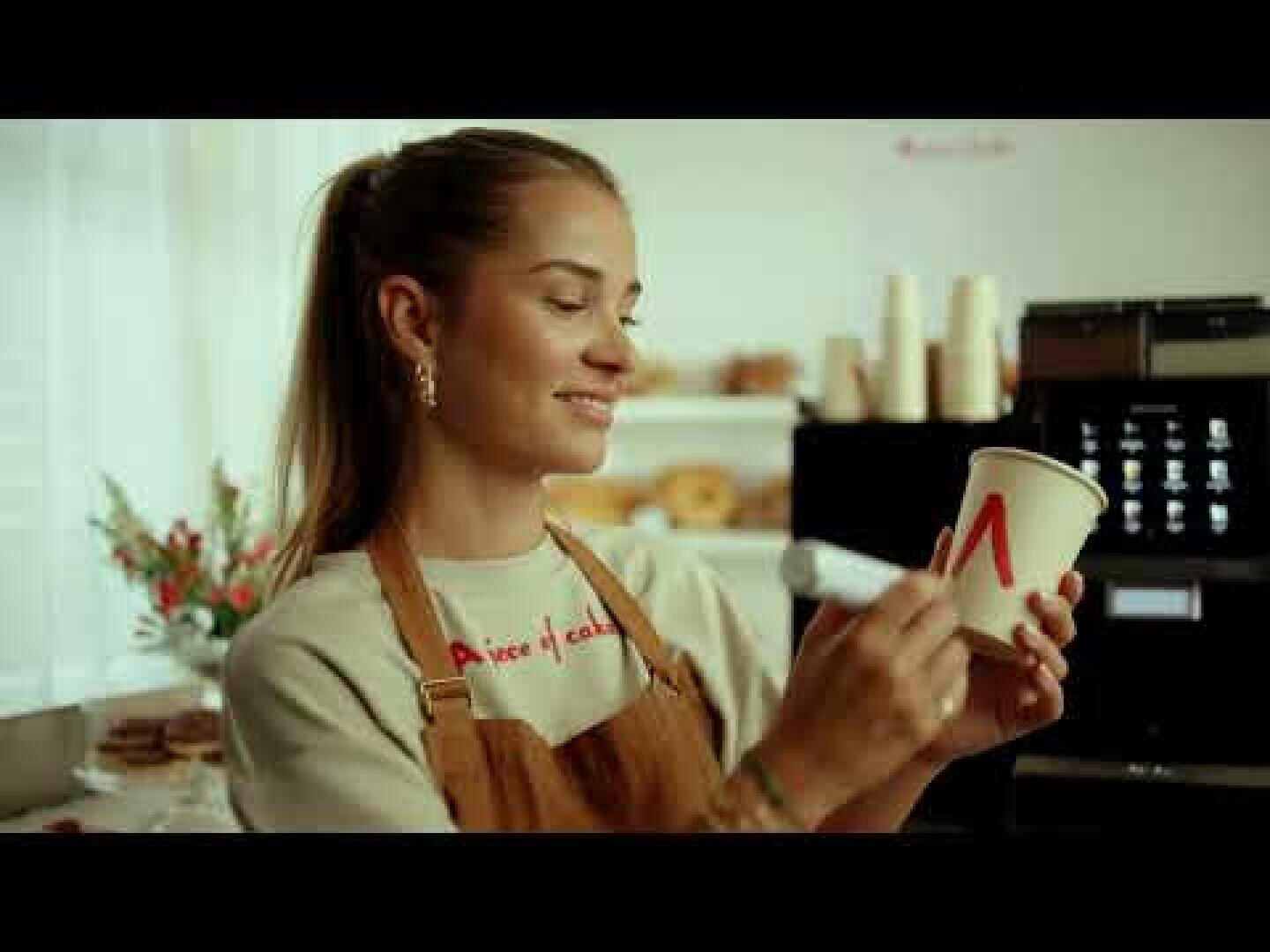 Ein lächelnder Barista in einer Schürze schreibt mit einem Marker den Buchstaben M auf einen Papierkaffeebecher in einem gemütlichen Café, in dem im Hintergrund Regale und Gebäck zu sehen sind.