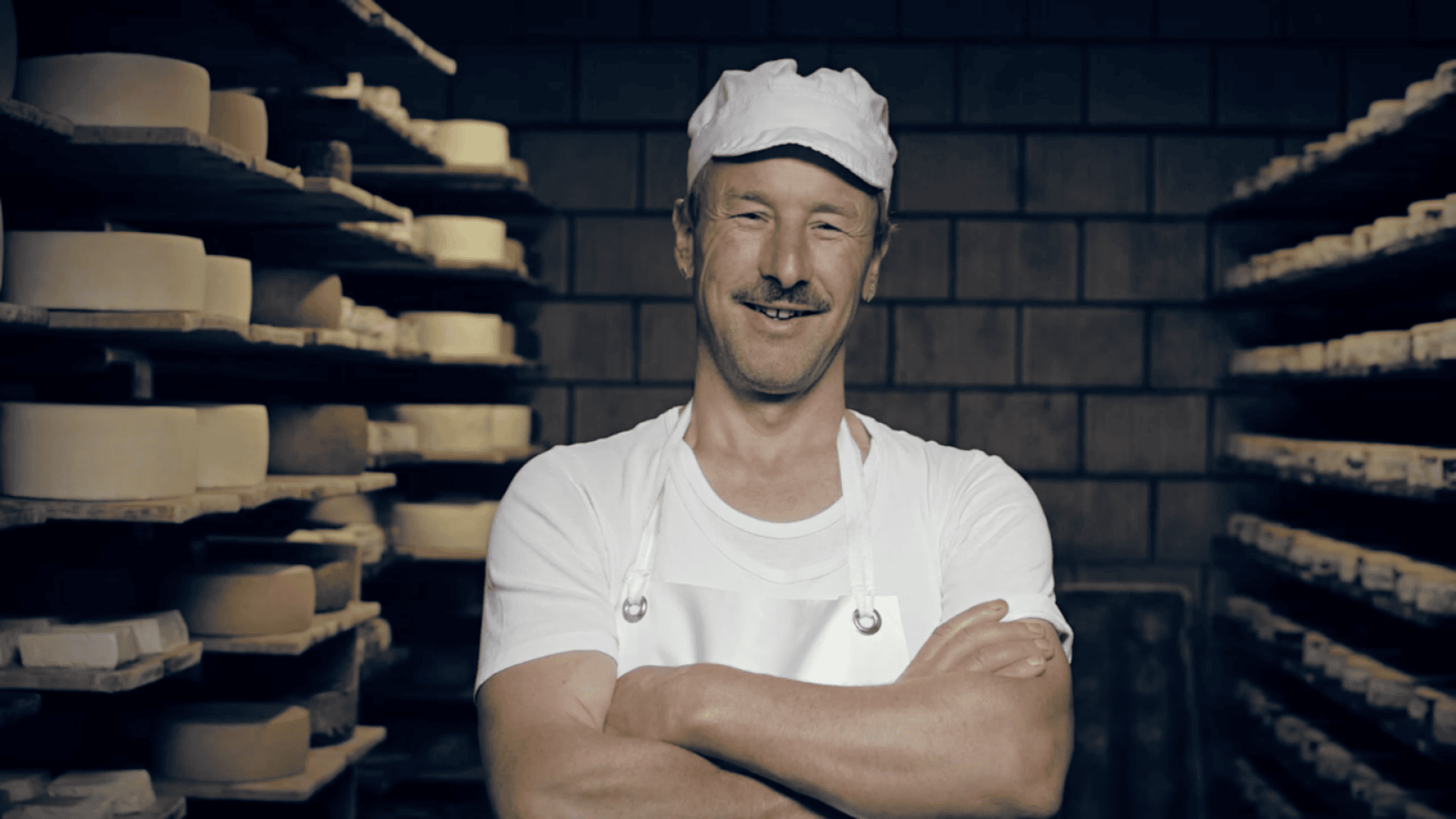 Ein lächelnder Mann in weißem Hemd, Schürze und Mütze steht mit verschränkten Armen vor Regalen voller Käselaibe in einem schwach beleuchteten Raum.