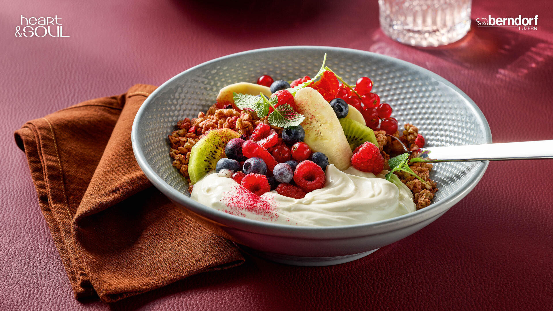Eine Schale Müsli, garniert mit Joghurt, Blaubeeren, Himbeeren, Johannisbeeren, Kiwischeiben, einer Banane und einem Minzblatt, steht auf einem roten Tisch mit einer braunen Serviette und einem Löffel. Im Hintergrund befindet sich ein Glas Wasser.