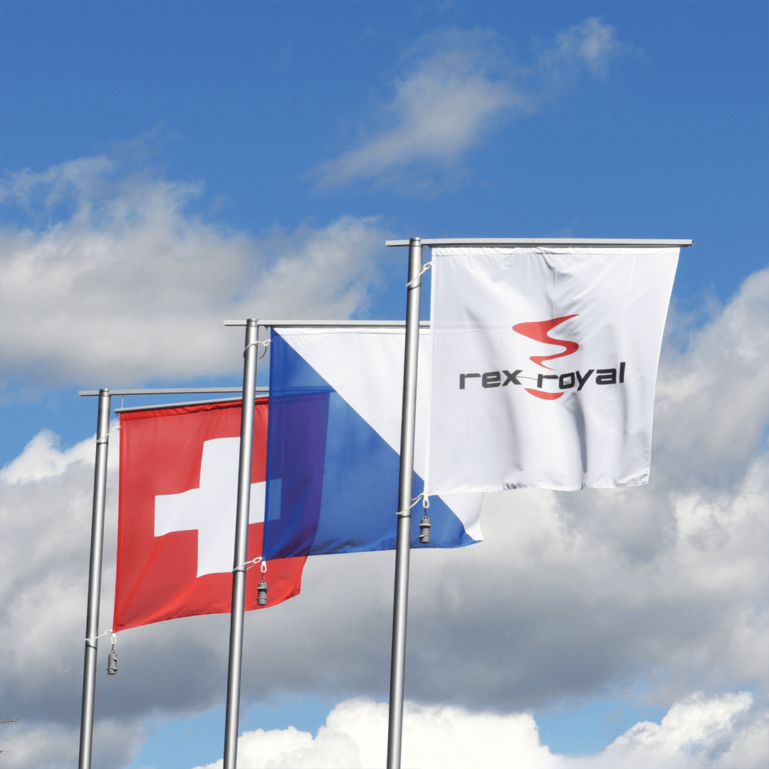 Trois drapeaux sur des mâts devant un ciel partiellement nuageux : le drapeau suisse (rouge avec une croix blanche), un drapeau bleu et blanc et un drapeau blanc avec le logo et le texte de rex-royal.