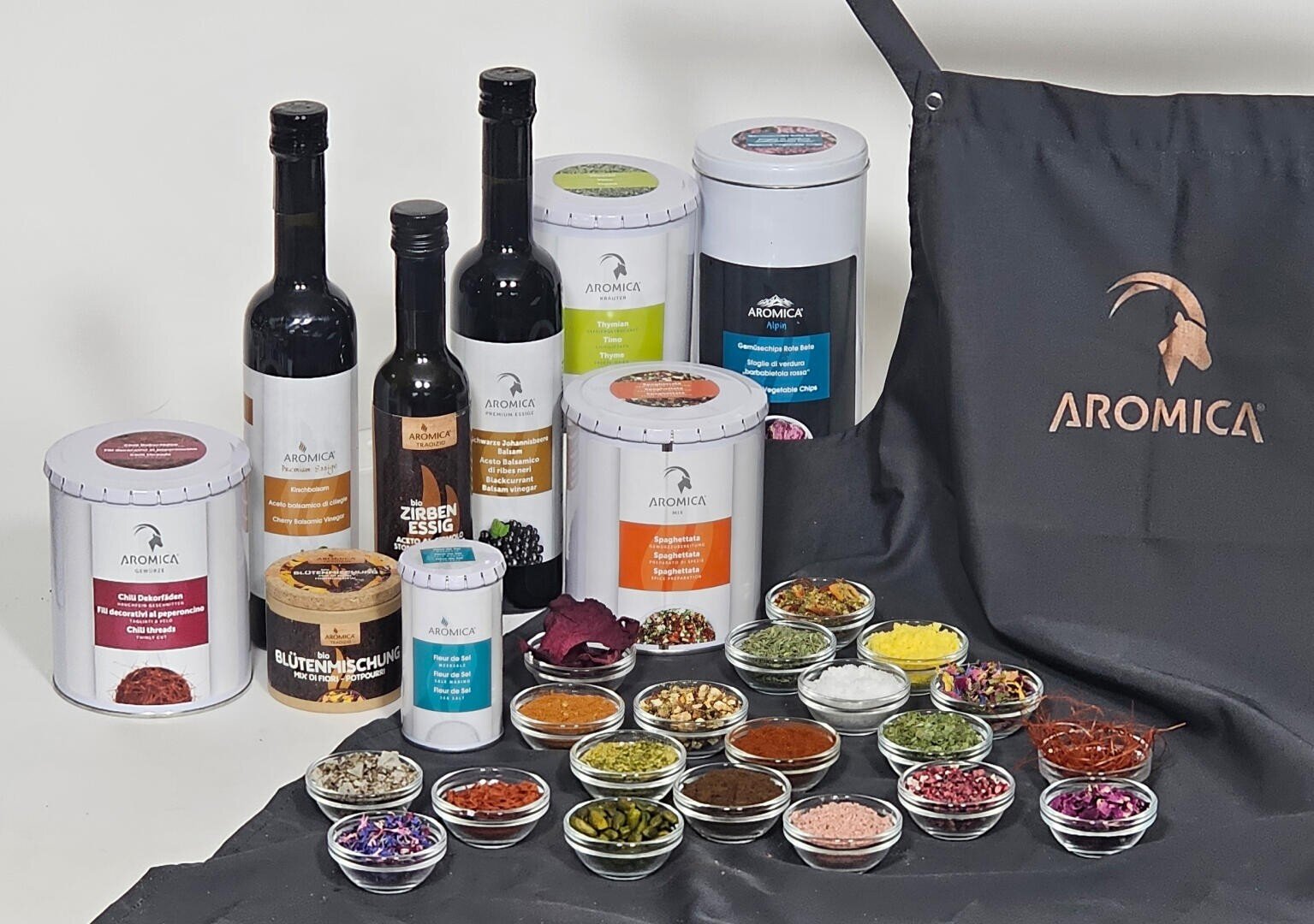 Une collection de produits alimentaires Aromica, comprenant des épices, des huiles et des vinaigres en bocaux et en bouteilles, disposés avec de petits bols d'épices colorées devant, et un tablier noir Aromica à droite.