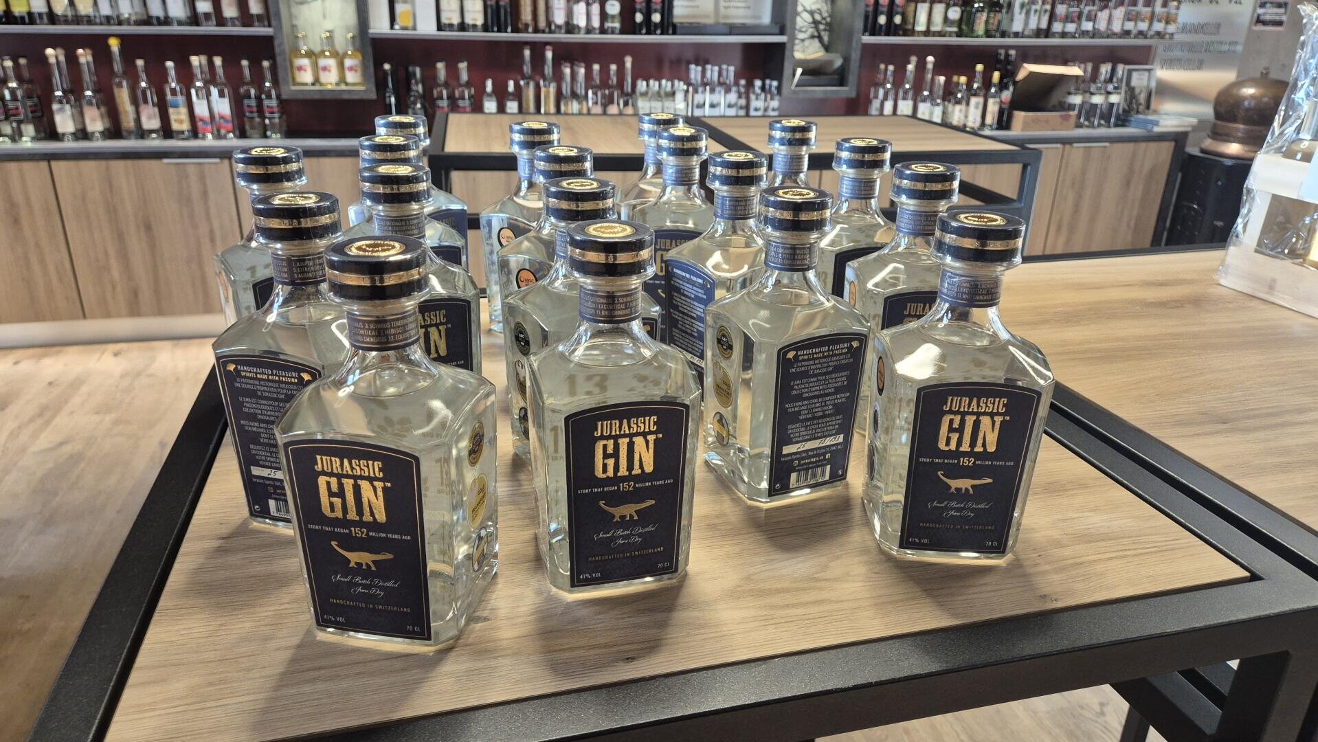 Mehrere Flaschen Jurassic Gin mit dunkelblauen Etiketten stehen auf einem hölzernen Auslagetisch in einem Spirituosengeschäft, während im Hintergrund Regale mit anderen Spirituosen zu sehen sind.