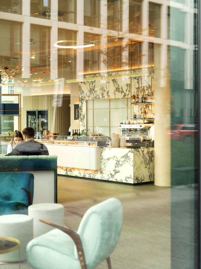 Ein modernes Café mit Marmortheken, goldenen Akzenten und mit Tassen und Flaschen bestückten Regalen. Zwei Personen sitzen an einem Tisch, während Plüschsessel und natürliches Licht eine gemütliche, einladende Atmosphäre schaffen. Die Szene ist durch große Fenster zu sehen.