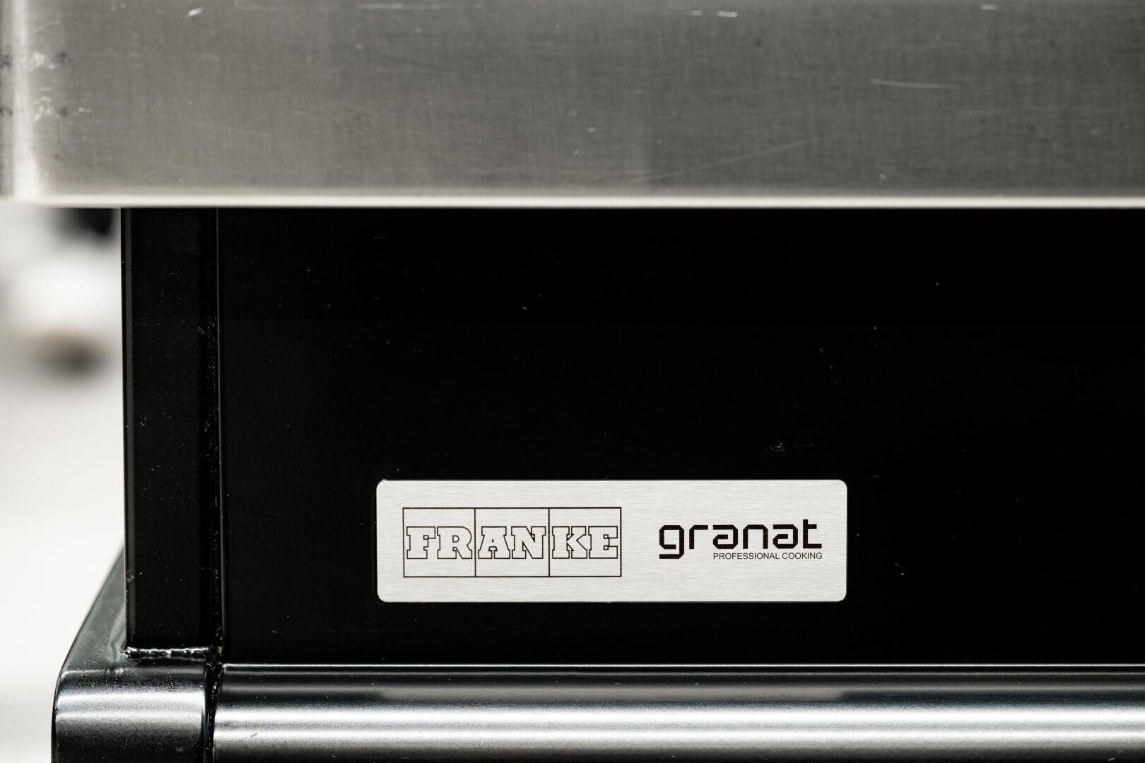 Nahaufnahme eines Metallschildes auf einem schwarzen Gerät mit den Logos FRANKE und granat PROFESSIONAL COOKING unter einer Edelstahloberfläche.