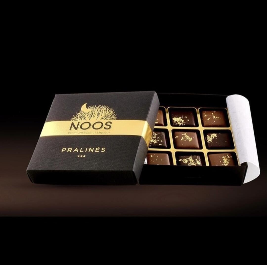 Eine schwarz-goldene Schachtel mit der Aufschrift NOOS Pralinés ist geöffnet und gibt den Blick auf neun fein säuberlich angeordnete Schokoladenpralinen frei, die jeweils mit Goldflocken bestreut sind und sich vor einem dunklen Hintergrund befinden.
