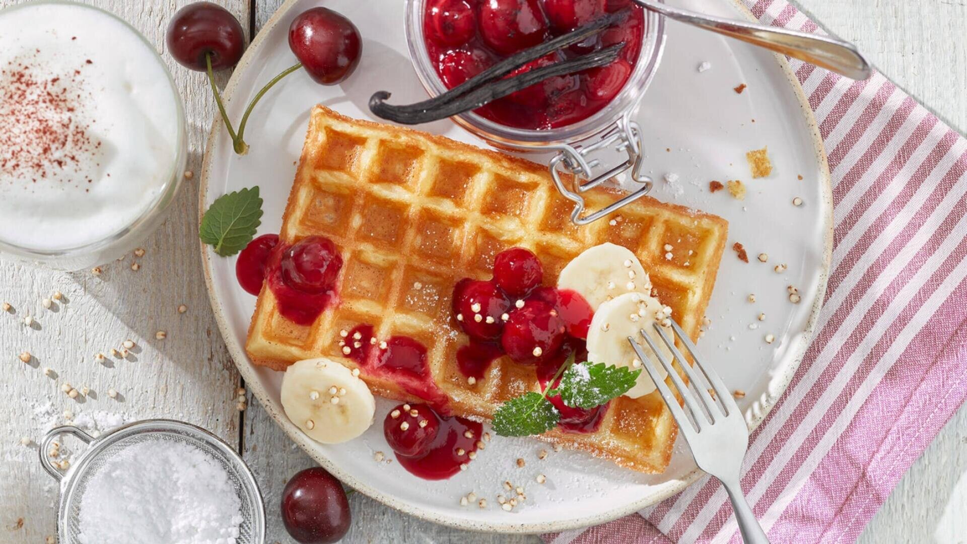 Ein Teller mit einer goldenen Waffel, die mit Bananenscheiben, Kirschtopping und Minzblättern belegt ist. Eine Gabel liegt auf dem Teller. Daneben stehen ein Glas mit aufgeschäumter Milch, frische Kirschen, ein Glas mit Kompott und eine gestreifte Serviette.