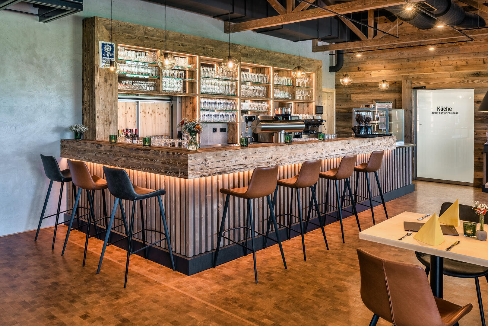 Un bar moderne avec un comptoir en bois, de hauts tabourets bruns, des étagères avec des verres et des bouteilles et un éclairage chaleureux. Sur le comptoir et les tables, il y a de petits vases de fleurs et, en arrière-plan, une porte avec l'inscription "cuisine".