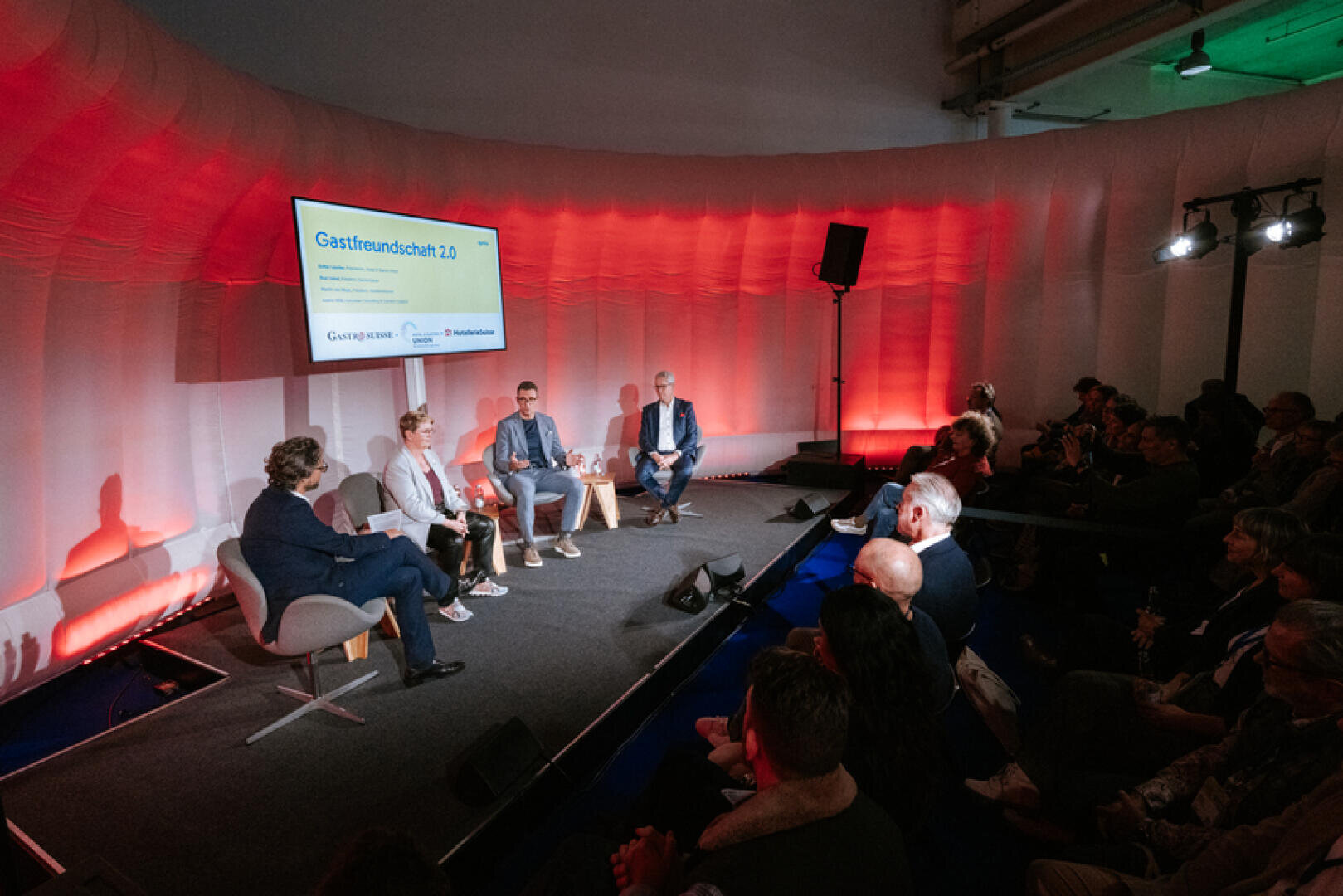 Eine Gruppe von vier Personen sitzt auf einer Bühne und diskutiert vor einem Publikum. Hinter ihnen wird auf einem Bildschirm "Gastfreundschaft 2.0" angezeigt, und die Kulisse wird rot beleuchtet. Die Veranstaltung scheint eine Konferenz oder ein Seminar zu sein.