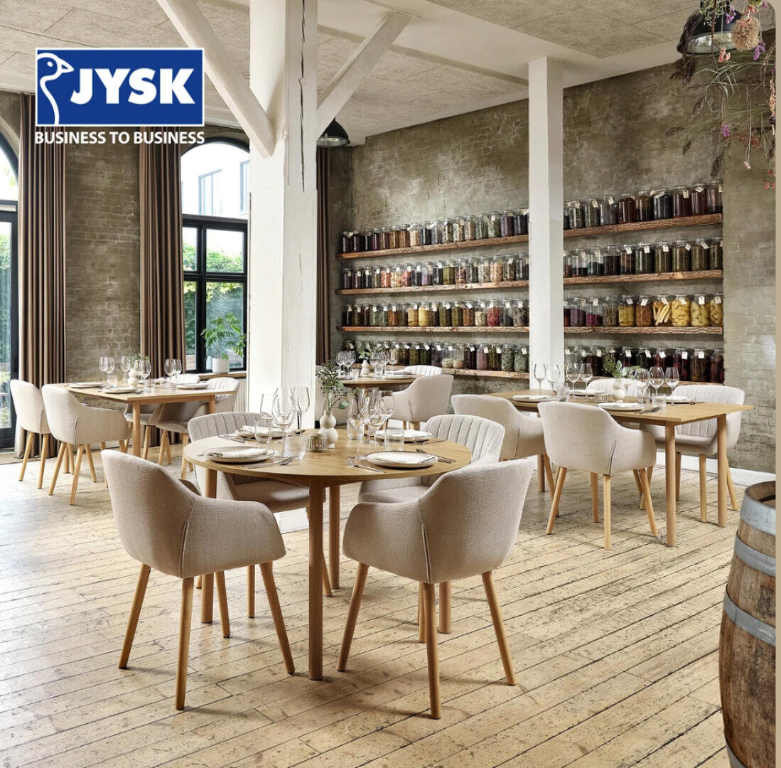 Modernes Restaurantinterieur mit runden Holztischen und gepolsterten Stühlen, die zum Essen aufgestellt sind. Gläser stehen in Regalen an einer rustikalen Backsteinwand. Große Fenster lassen natürliches Licht herein. JYSK Business to Business-Logo in der oberen linken Ecke.
