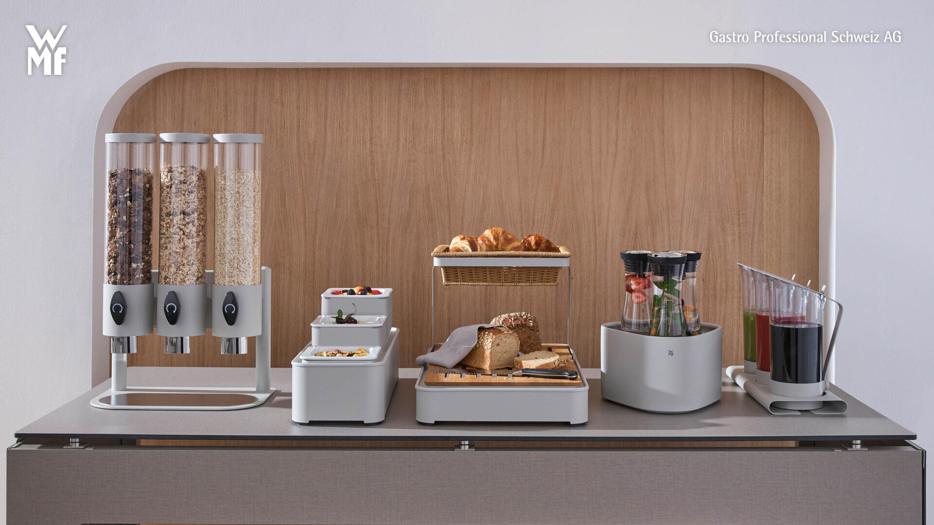 Un buffet de petit-déjeuner avec des distributeurs de céréales, des récipients contenant des fruits et des yaourts, une corbeille de croissants, des tranches de pain, un pichet de jus de fruits et des verres avec des ustensiles sur un plan de travail moderne.