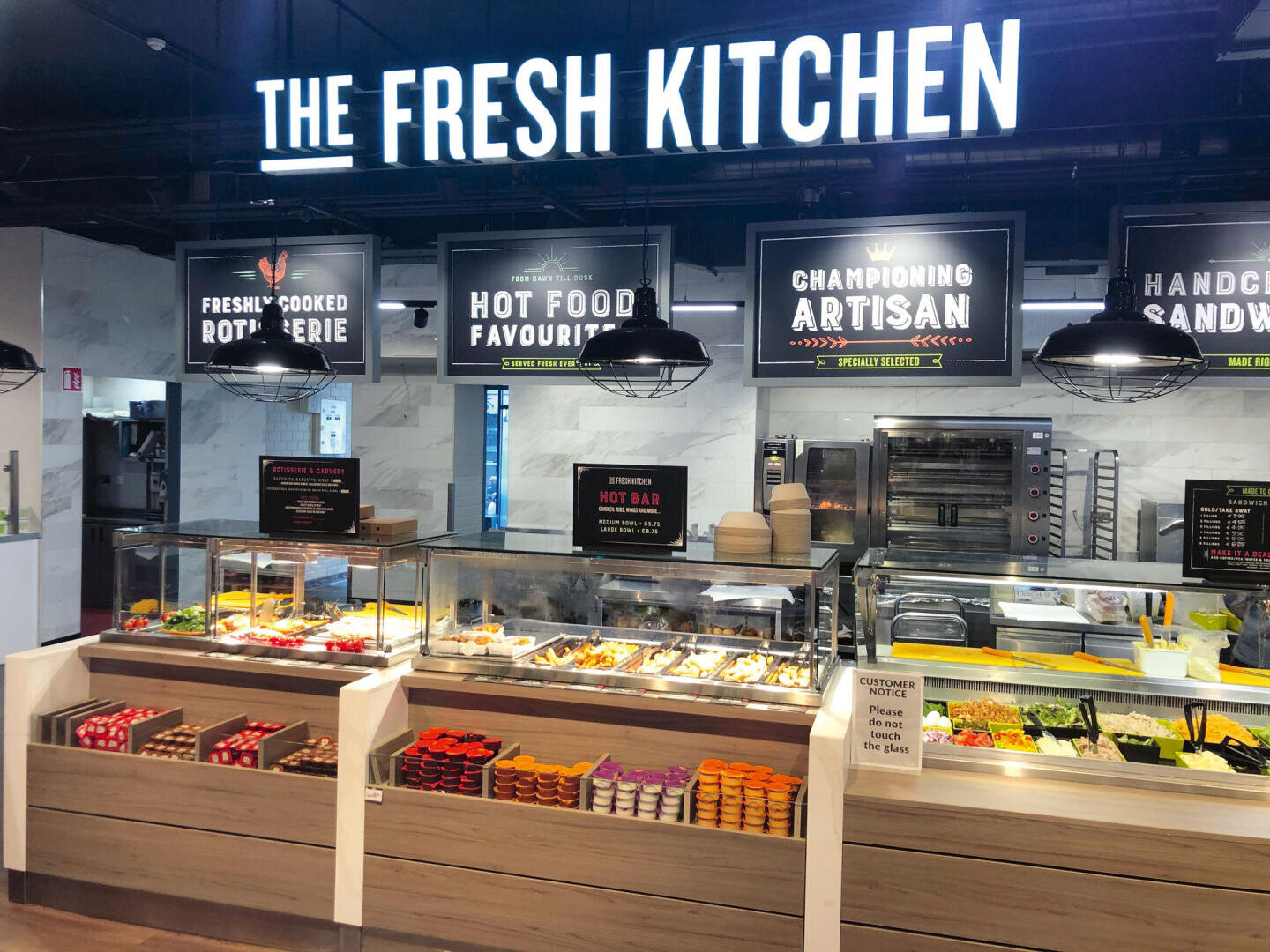 Un comptoir moderne en libre-service, appelé "The Fresh Kitchen", propose une grande variété de plats chauds et froids, de salades, de fruits et d'aliments emballés dans un environnement propre, bien éclairé et sous une signalétique claire.