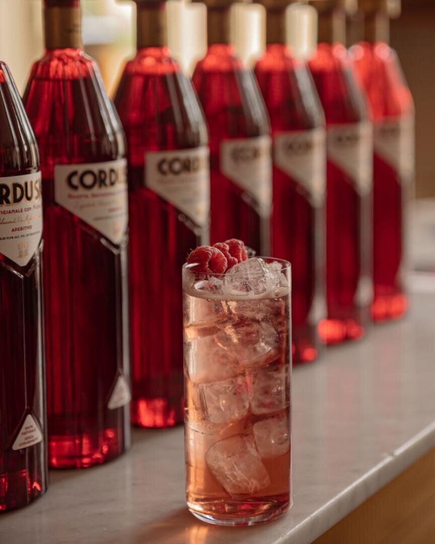Ein mit Eis gefülltes Glas mit einem roten Getränk und Himbeeren darauf steht auf dem Tresen vor einer Reihe roter Cordus-Flaschen.
