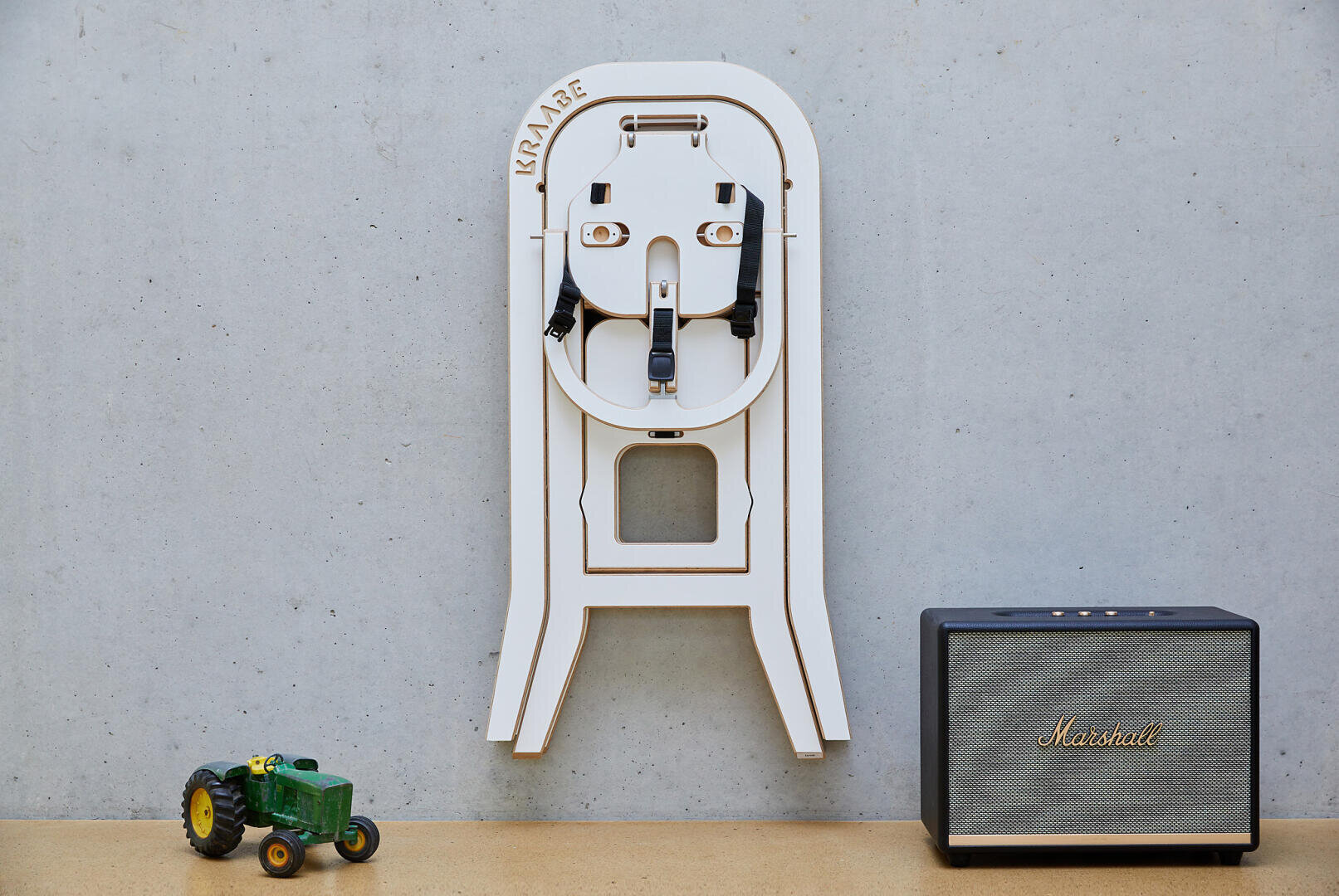 Ein weißer klappbarer Hochstuhl ist an einer grauen Wand montiert. Darunter befinden sich ein kleiner grüner Spielzeugtraktor und ein schwarzer Marshall-Lautsprecher auf einer hellen Holzfläche.