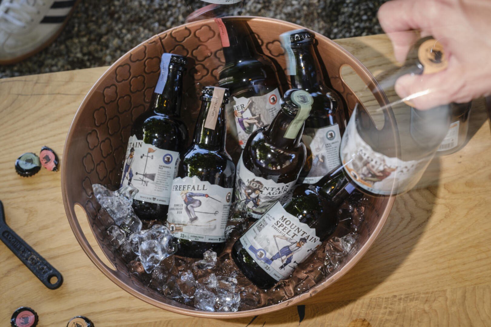 Ein mit Eis gefüllter Kupfereimer und mehrere Bierflaschen stehen auf einem Holztisch. Eine Hand greift nach einer Flasche. Flaschendeckel und ein Flaschenöffner liegen in der Nähe verstreut. In der oberen linken Ecke ist ein Turnschuh teilweise sichtbar.