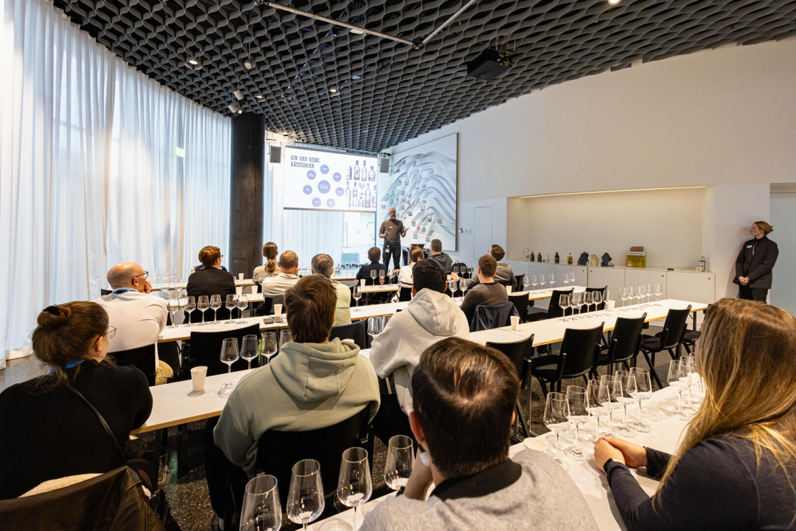 Eine Gruppe von Personen nimmt in einem modernen Raum an einer Weinverkostung teil und hört einem Dozenten zu, der mit Gesten Dias zum Thema Wein vorführt. Die Weingläser sind auf den Tischen vor jedem Teilnehmer angeordnet.