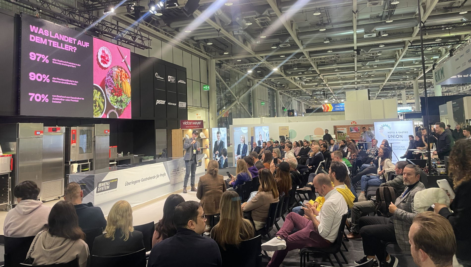 Ein Redner hält einen Vortrag vor einem sitzenden Publikum auf einer modernen Messe oder Konferenz. Auf einem großen Bildschirm werden Statistiken auf Deutsch mit einem Bild von Lebensmitteln angezeigt. Der Veranstaltungsort ist mit heller Beleuchtung, Bannern und Messeständen im Hintergrund ausgestattet.