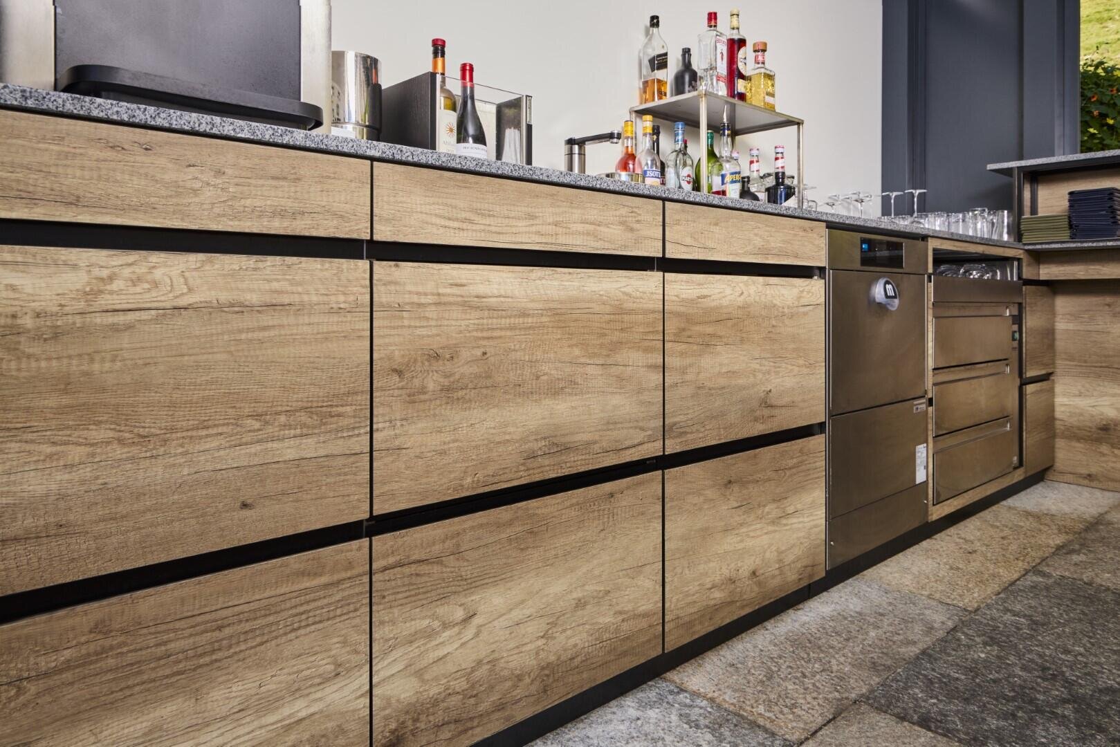 Unterer Blickwinkel auf einen modernen Barbereich mit Holzschränken, einem Geschirrspüler und einer Granitarbeitsplatte. Alkoholflaschen, Gläser und Utensilien sind ordentlich auf dem Tresen und in den Regalen angeordnet.