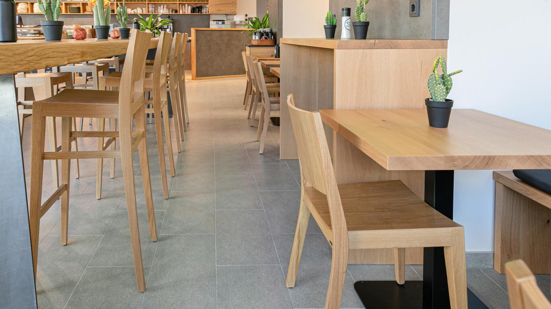 Intérieur d'un café moderne avec des tables et des chaises en bois clair, de petits cactus en pot sur les tables et un sol carrelé aux tons neutres. Des étagères contenant des plantes et des objets de décoration sont visibles à l'arrière-plan.
