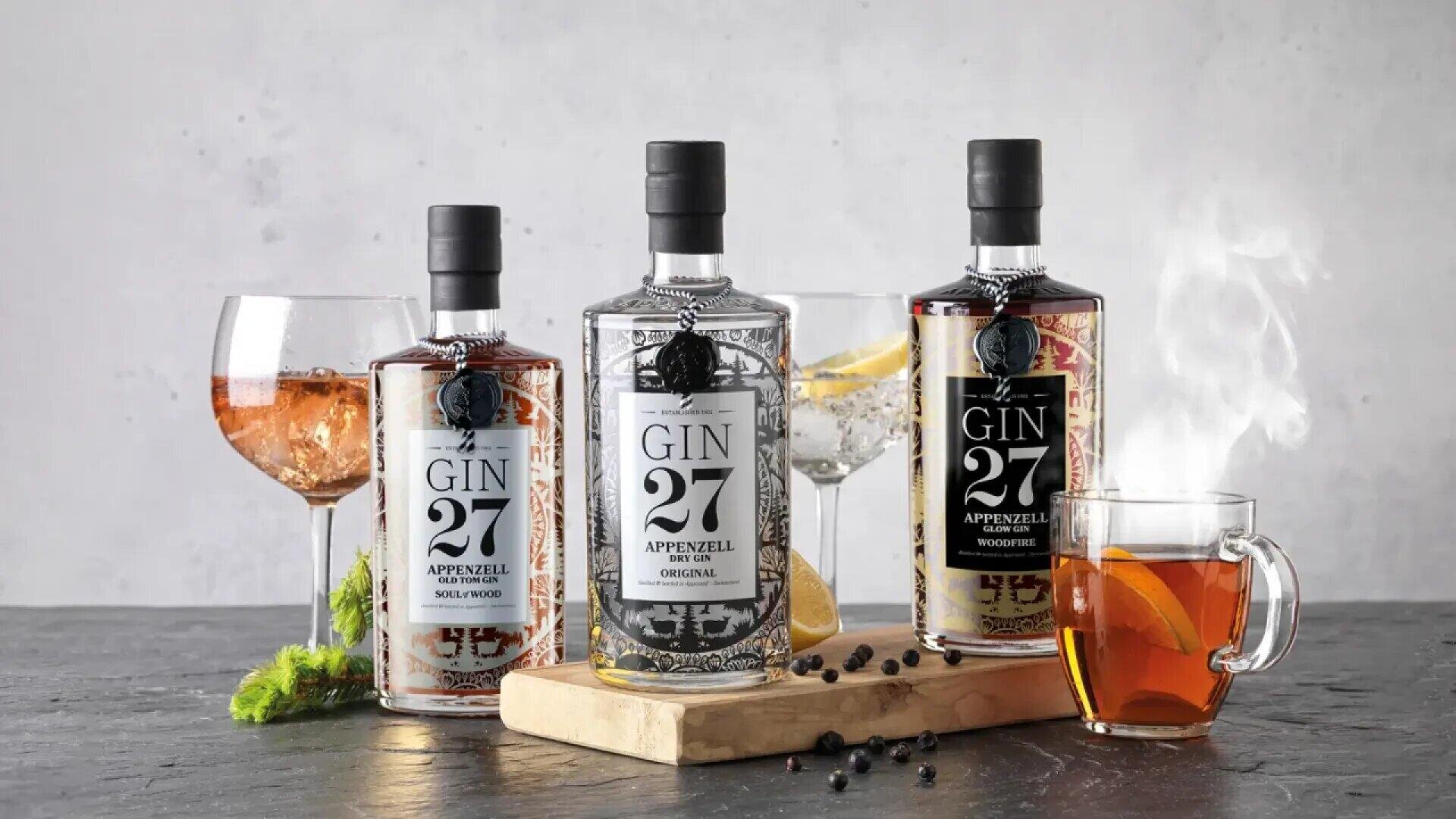Drei Flaschen Gin 27 Appenzell mit drei Getränken: ein G&T garniert mit Zitrone, ein Cocktail mit Kräutern und eine dampfende Tasse Tee, alles arrangiert auf einer hölzernen Oberfläche mit verstreuten Botanicals.