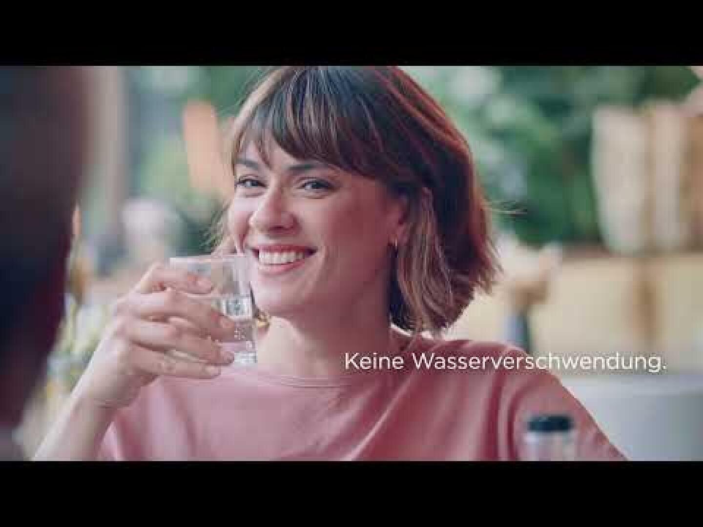 Eine Frau mit kurzen braunen Haaren lächelt, während sie ein Glas Wasser hält. Der Text auf dem Bild lautet: "Keine Wasserverschwendung". Der Hintergrund ist leicht unscharf und suggeriert eine Umgebung im Freien.