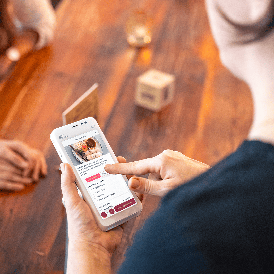 Une personne tenant un smartphone à la main et utilisant une application de commande de repas à une table en bois, les mains d'une autre personne étant visibles au-dessus de la table. L'écran du téléphone affiche un plat et des options de commande.