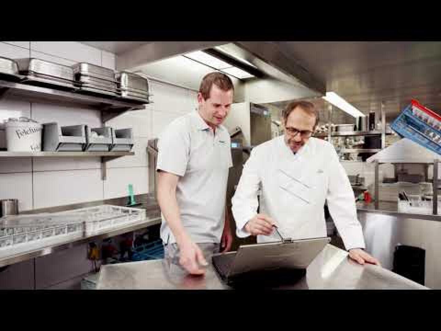 Deux hommes se tiennent dans une cuisine industrielle, l'un portant une veste de cuisine blanche, l'autre un polo gris. Ils regardent un ordinateur portable posé sur un comptoir en inox et semblent discuter de quelque chose sur l'écran.