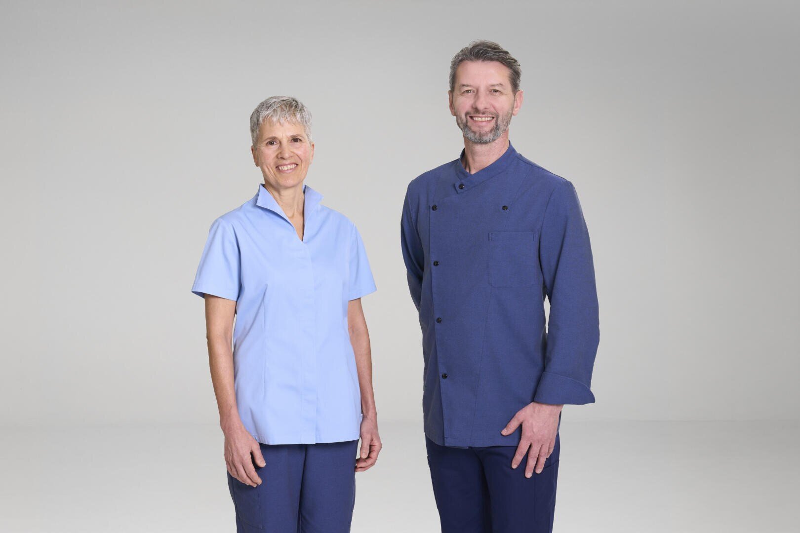 Zwei Erwachsene, einer in einem hellblauen kurzärmeligen Hemd und einer dunklen Hose, der andere in einem dunkelblauen Hemd mit Knöpfen und einer Hose, stehen nebeneinander und lächeln in einer schlichten, hellen Studioumgebung.