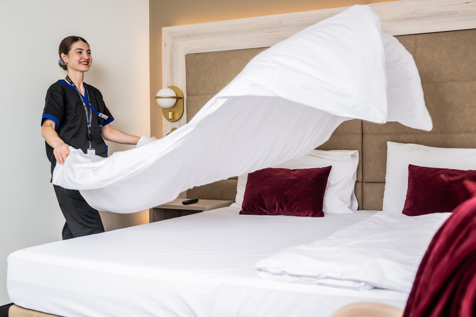 Eine Hotelangestellte lächelt, während sie ein Bett macht und ein weißes Laken über ein modernes, ordentlich eingerichtetes Zimmer mit roten Kissen wirft.