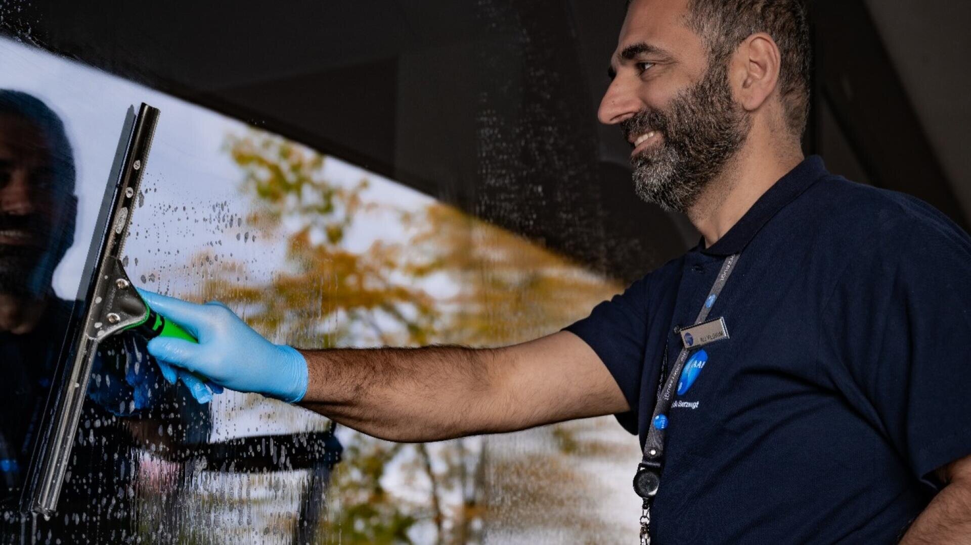 Ein Mann in einem marineblauen Hemd und blauen Handschuhen lächelt, während er ein Glasfenster mit einem Abzieher reinigt. Durch das nasse Glas sind Bäume und der Himmel zu sehen.