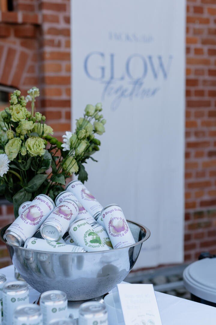 Eine silberne Schale, gefüllt mit verschiedenen Dosen, steht auf einem Tisch neben einem Strauß grüner und weißer Blumen. Im Hintergrund hängt ein weißes Banner mit der Aufschrift GLOW together an einer Backsteinwand.