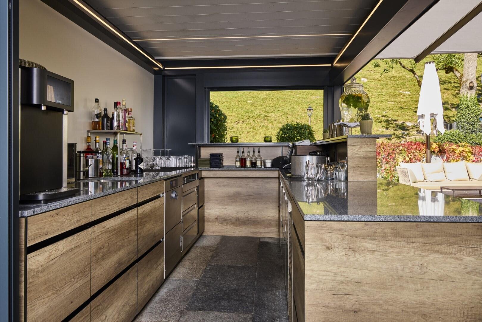 Moderne Bar im Freien mit Holzschränken, Granitarbeitsplatten, verschiedenen Flaschen und Gläsern, einer Kaffeemaschine und einem Sitzbereich, der durch ein großes, offenes Fenster zum Garten hin sichtbar ist.