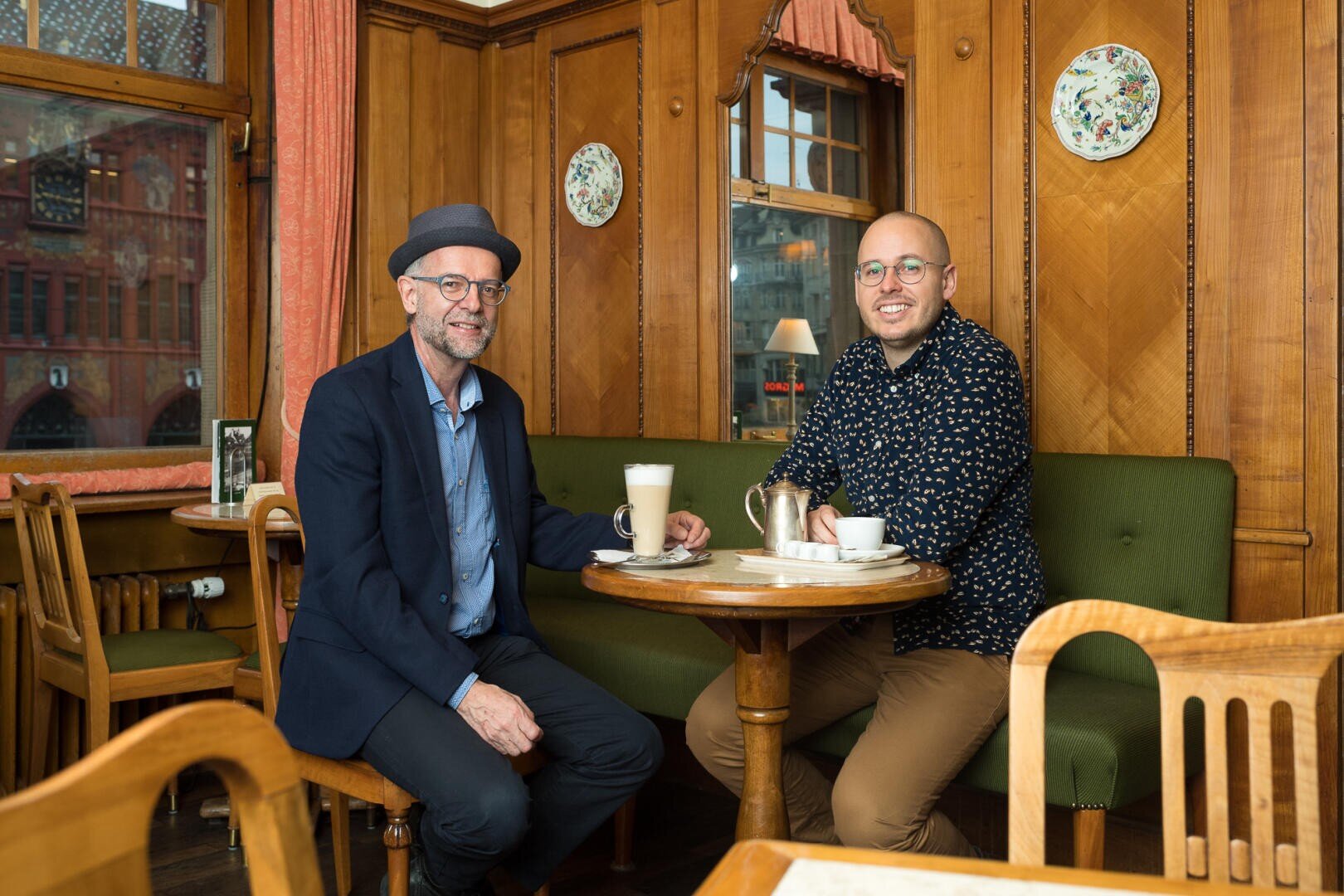 Zwei Männer sitzen an einem kleinen runden Tisch in einem gemütlichen, holzgetäfelten Café und lächeln. Der eine trägt einen Hut und eine Brille, der andere eine Brille und ein gemustertes Hemd. Vor ihnen stehen Kaffeegetränke. Zur Dekoration gehören Teller und große Fenster.