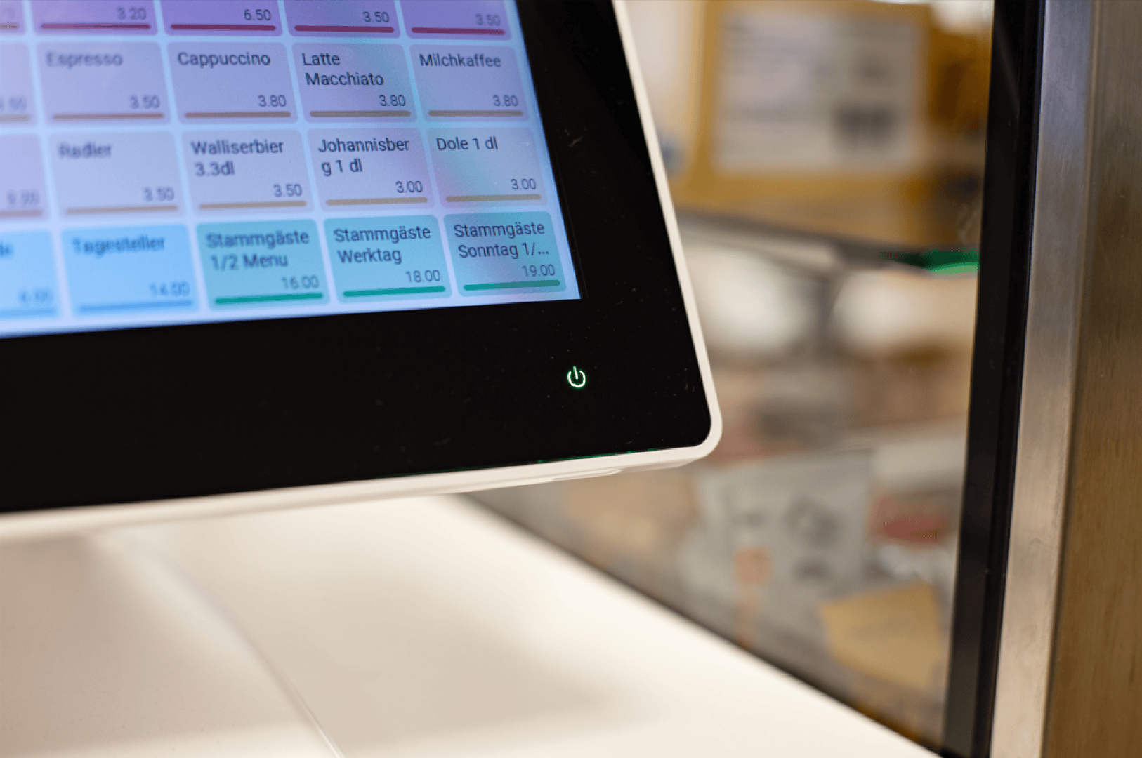 Eine Nahaufnahme eines Touchscreen-Menüs mit Getränkeoptionen auf Deutsch, darunter Espresso und Latte Macchiato. Auf dem Gerät ist ein grüner Einschaltknopf mit einem unscharfen Hintergrund auf der rechten Seite sichtbar.