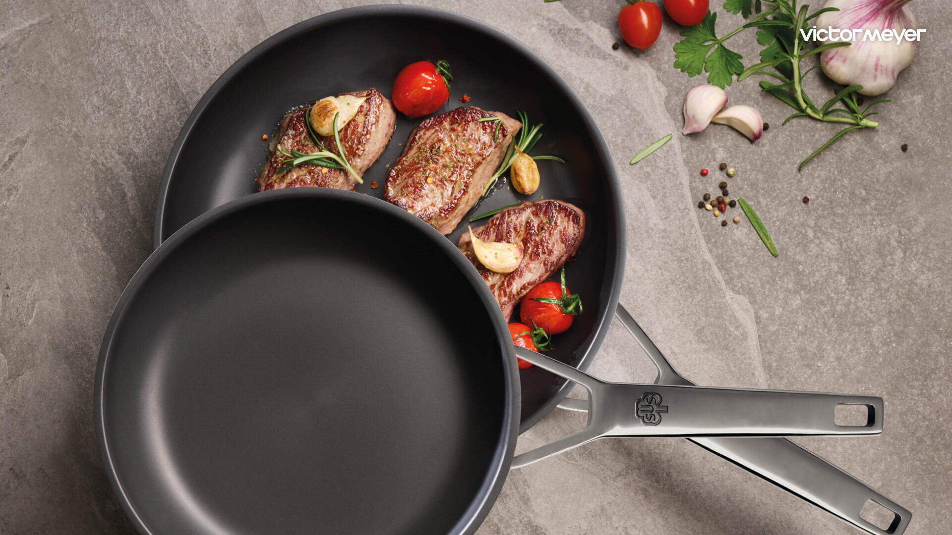 Zwei Bratpfannen aus rostfreiem Stahl auf einem Küchentisch; eine Pfanne enthält angebratenes Fleisch, Kirschtomaten, Knoblauch und Kräuter. Die andere Pfanne ist leer. Frische Zutaten und Pfefferkörner liegen in der Nähe verstreut.