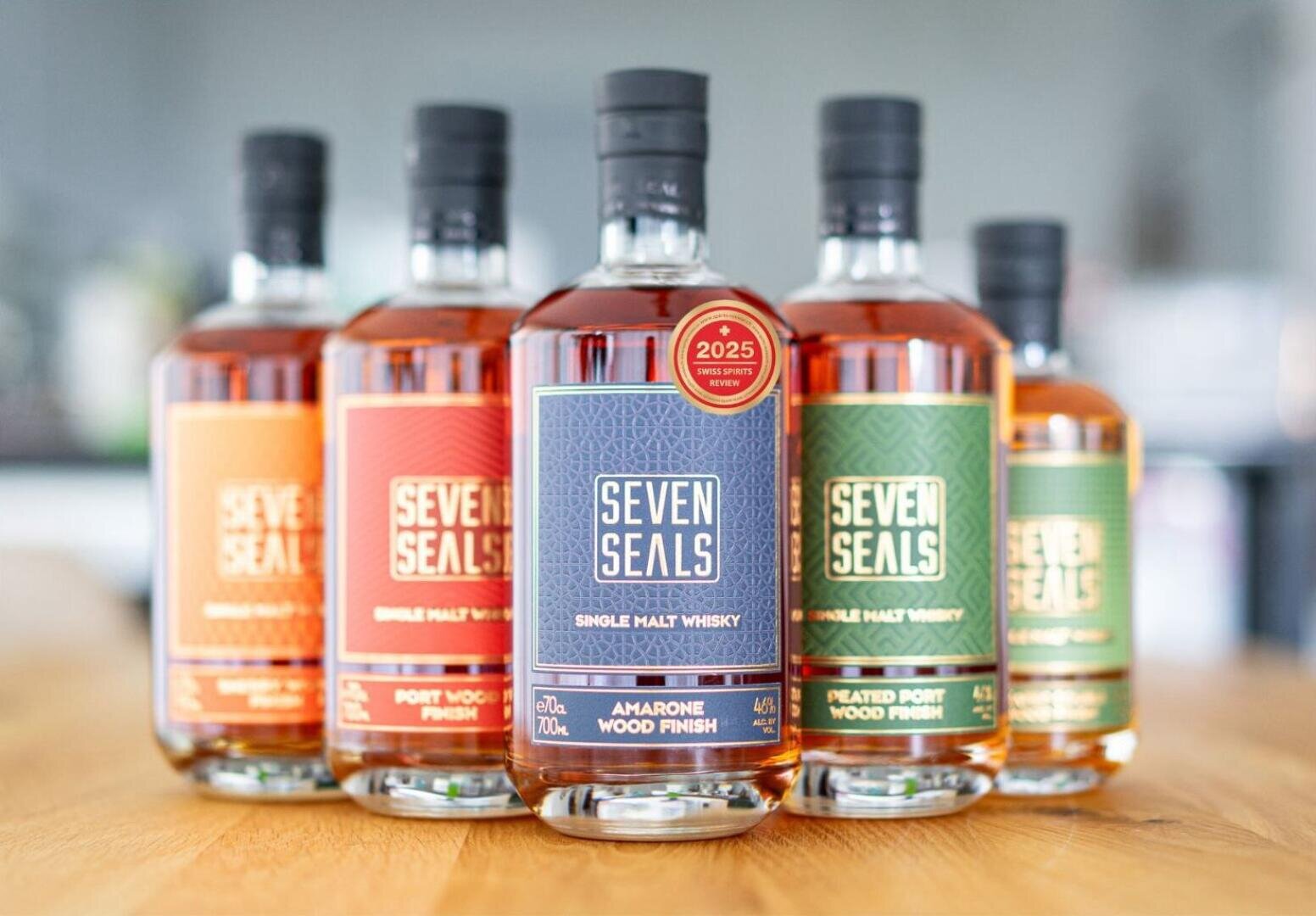 Fünf Flaschen Seven Seals Single Malt Whisky in verschiedenen Etikettenfarben sind auf einer Holzfläche angeordnet, wobei die mittlere Flasche mit der Aufschrift Amarone Wood Finish im Mittelpunkt steht. Der Hintergrund ist sanft verschwommen.