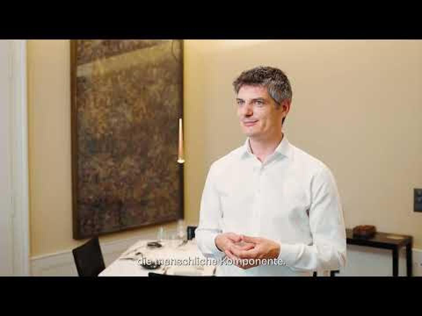 Ein Mann in einem weißen Hemd steht in einem Raum, lächelt und gestikuliert mit den Händen. Er befindet sich in einem gut beleuchteten Raum mit einem großen abstrakten Gemälde an der Wand und einem gedeckten Esstisch im Hintergrund. Am unteren Rand erscheinen Untertitel.